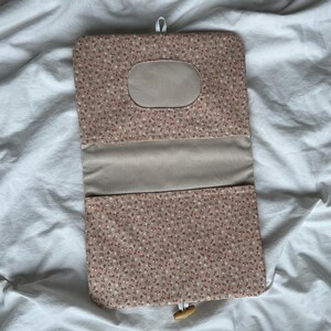 May include: A rectangular fabric pouch featuring a small-scale floral pattern in shades of pink, white, and green. The pouch has a solid beige interior panel, a central oval cutout, and a wooden toggle closure attached to a white elastic loop.
