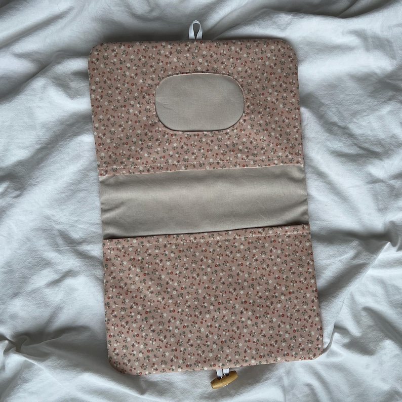 May include: A rectangular fabric pouch featuring a small-scale floral pattern in shades of pink, white, and green. The pouch has a solid beige interior panel, a central oval cutout, and a wooden toggle closure attached to a white elastic loop.