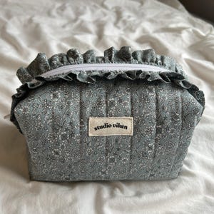 May include: A small, quilted cosmetic bag in a floral pattern of white and gray on a blue-green background. The bag has a ruffled trim and a white zipper. The label reads "studio viken".