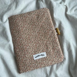 May include: A quilted fabric sleeve in a dusty pink hue featuring a small floral pattern with white and red blossoms. The sleeve has diagonal stitching, a wooden toggle closure on the side, and a white rectangular label reading studio viken.