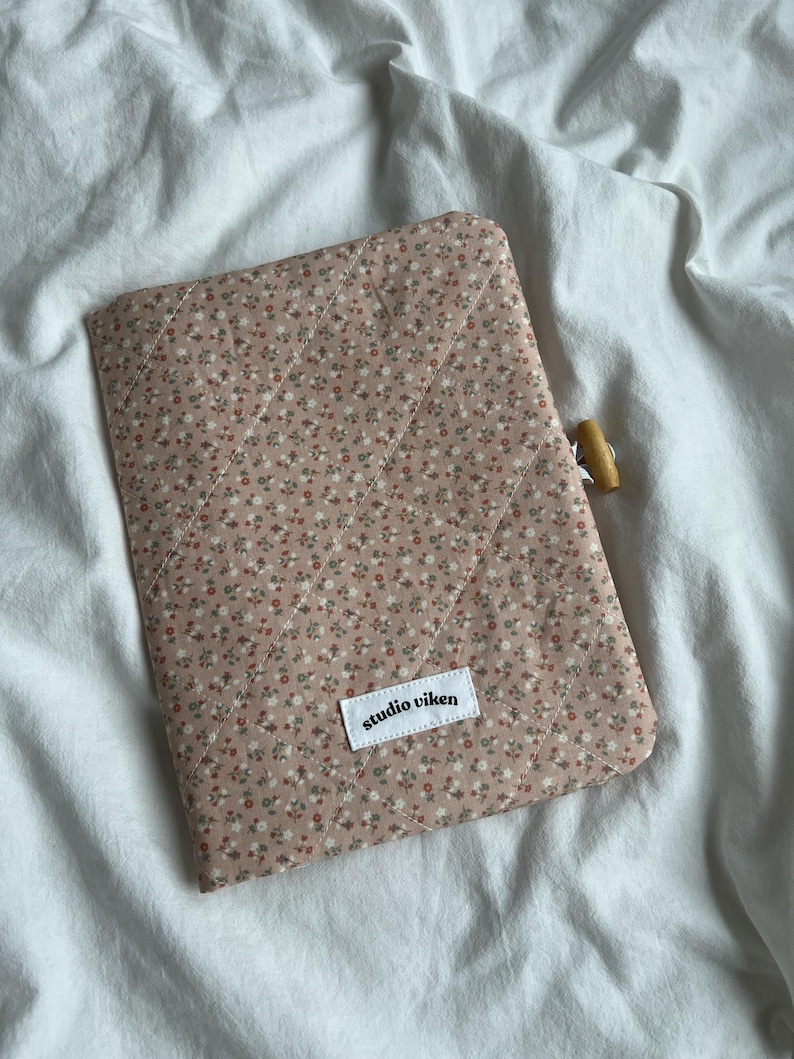 May include: A quilted fabric sleeve in a dusty pink hue featuring a small floral pattern with white and red blossoms. The sleeve has diagonal stitching, a wooden toggle closure on the side, and a white rectangular label reading studio viken.