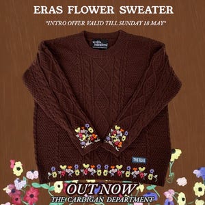 May include: A dark brown cable-knit sweater with floral embroidery on the cuffs and hem. The text "ERAS FLOWER SWEATER" and "OUT NOW" are displayed. The sweater has a crew neck and a label that says "we will be remembered".