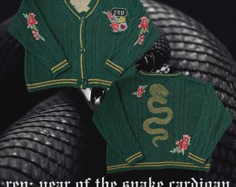May include: Green cable-knit cardigan with gold trim and floral embroidery. Features a snake design on the back and the text "rep: year of the snake cardigan" and "THE CARDIGAN DEPARTMENT".