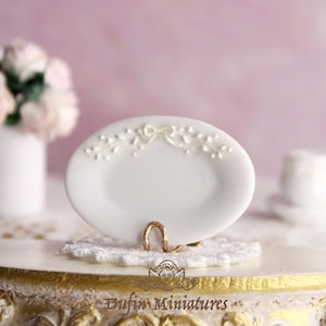 Può includere: Un piatto in porcellana bianca in miniatura con un delicato motivo floreale e un nastro bianco. Il piatto è su un pizzo bianco e un supporto dorato.