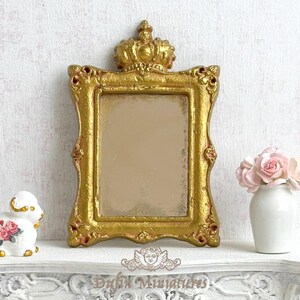 Pode incluir: Um espelho dourado adornado com uma coroa no topo. O espelho está em uma moldura decorativa com acabamento dourado. O espelho está em uma prateleira branca com uma figura de cerâmica branca e um vaso branco com flores cor-de-rosa.