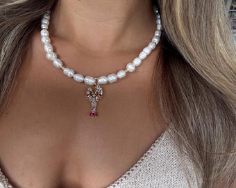 May include: A white pearl necklace with a gold lobster pendant. The lobster pendant is encrusted with colorful gemstones, including red, blue, and yellow. The necklace is worn around the neck.