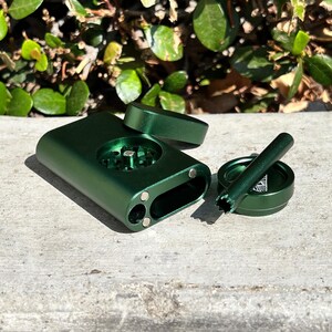 May include: A disassembled, metallic green grinder set. The set includes a rectangular main body, a circular top, and a small cylindrical tool. The main body has a circular grinding chamber and two small holes. The circular top has a logo. The set is on a concrete surface.