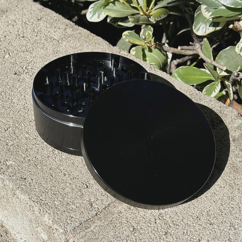 May include: A black, cylindrical herb grinder with a lid. The grinder is made of metal and has sharp teeth inside. The lid is off to the side, revealing the grinding surface. The grinder is sitting on a concrete surface.