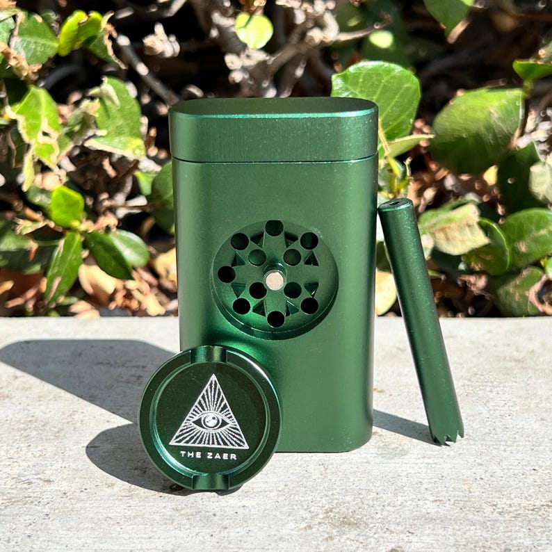 May include: A green metal grinder set with a rectangular grinder, a small cylindrical tool, and a round ashtray. The ashtray features a white triangle with an eye and the text "THE ZAER". The grinder has a circular design.