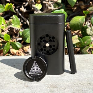 May include: A black metal grinder with a circular grinding chamber and a matching black ashtray. The ashtray features a white eye-in-triangle design and the text "THE ZAER". A black poker tool is also included.