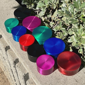 May include: A collection of colorful, cylindrical grinders in various shades, including black, green, blue, red, and pink. The grinders are arranged on a concrete surface with green foliage in the background. Each grinder has a circular top.