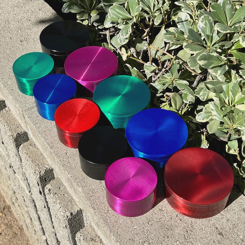 May include: A collection of colorful, cylindrical grinders in various shades, including black, green, blue, red, and pink. The grinders are arranged on a concrete surface with green foliage in the background. Each grinder has a circular top.