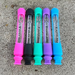 Puede incluir: Cinco chillums de vidrio de colores con fundas de silicona. Los chillums son de color rosa, negro, verde azulado, morado y azul. Cada uno tiene el texto "THE ZAER & CHILLUM" impreso.