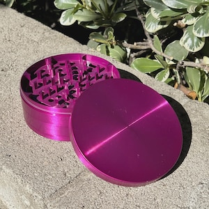 May include: A magenta-colored, two-piece grinder with a circular design. The top piece is a solid disc, while the bottom piece has a textured interior. The grinder is made of metal and sits on a concrete surface.