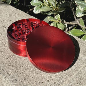 May include: A red, two-piece herb grinder with a textured interior and a smooth, circular lid. The grinder is made of metal and sits on a concrete surface, with green foliage in the background.