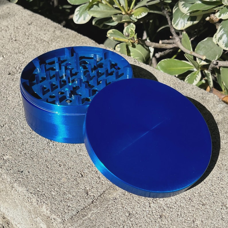 May include: A blue grinder with a lid, designed for herbs or spices. The grinder is made of metal and has a textured interior. The lid is a solid, smooth blue. The grinder is sitting on a concrete surface.