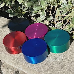 May include: A collection of five round, metallic grinders in various colors: red, blue, green, pink, and black. Each grinder has a brushed metal finish and a cylindrical shape, with a flat top and bottom. The grinders are arranged on a stone surface with green foliage in the background.