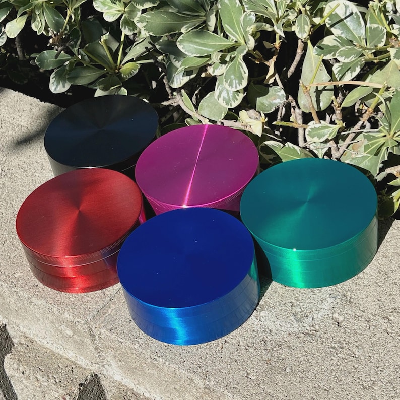 May include: A collection of five round, metallic grinders in various colors: red, blue, green, pink, and black. Each grinder has a brushed metal finish and a cylindrical shape, with a flat top and bottom. The grinders are arranged on a stone surface with green foliage in the background.