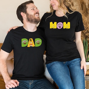 May include: Black t-shirts with Sesame Street character designs. One shirt says "DAD" with Bert in the center and Ernie on either side. The other shirt says "MOM" with Big Bird and a purple background.