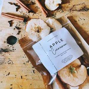 May include: A clear bag of apple and cinnamon simmer pot blend with dried apple slices and cinnamon sticks. The label reads "Apple + Cinnamon" and "Michigan Crafted". The ingredients are infused with spicy cloves and sweet cinnamon.