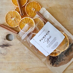 May include: Dried orange slices and cinnamon sticks are arranged on a wooden surface. A clear bag contains a simmer pot blend with the text "Pumpkin + Spice". The blend includes orange, cinnamon, and cloves, perfect for a warm, inviting scent.