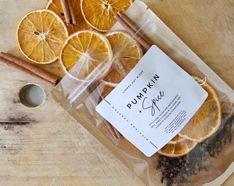 May include: Dried orange slices and cinnamon sticks are arranged on a wooden surface. A clear bag contains a simmer pot blend with the text "Pumpkin + Spice". The blend includes orange, cinnamon, and cloves, perfect for a warm, inviting scent.