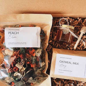 May include: A gift box containing a potpourri bag labeled "Peach Blossom", a jar of honey with a wooden honey dipper, and an "Oatmeal, Milk & Honey" artisan soap bar. The box is filled with brown paper shreds.