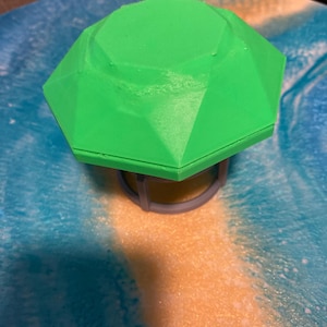 May include: A vibrant green, geometric object with a faceted top and a grey, circular base. The object appears 3D-printed and rests on a surface with blue, white, and orange tones.