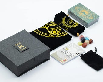 May include: A tarot card set with a black box featuring the text "1909". The set includes a deck of cards, a guide book, black velvet bags with gold designs, and a collection of colorful stones. The cards have a teal and gold design.
