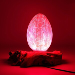 Puede incluir: Lámpara ovoide brillante con un patrón blanco agrietado, que emite luz roja y rosa. Descansa sobre un trozo de madera natural y oscura, sobre un fondo rojo. Un cable de alimentación negro se extiende desde la base.