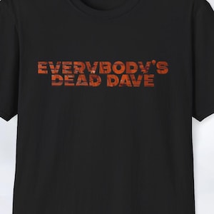 May include: Black crew neck t-shirt featuring the text "EVERYBODY'S DEAD DAVE" in an orange, brick-like font. The short-sleeved shirt is a casual style. The design is centrally placed on the chest.