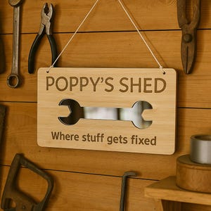 Pode incluir: Uma placa de madeira com o texto "POPPY'S SHED" e a frase "Where stuff gets fixed". A placa tem um recorte em forma de chave inglesa. Está pendurada numa parede de madeira com ferramentas.