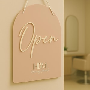 Op de afbeelding: Een roze, gebogen "Open" bord met de tekst "Open" in een cursief lettertype. Het bord bevat ook de tekst "HBM Hair by Maddi" in een kleiner, cursief lettertype. Het bord hangt aan een crèmekleurig touwtje.