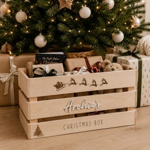 May include: A light-coloured wooden crate featuring a laser-engraved silhouette of Santa in a sleigh pulled by reindeer on the top slat. The middle slat displays the white 3D text "Archie's" and the bottom slat reads "CHRISTMAS BOX" with a tree icon.