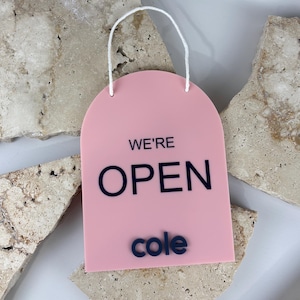 May include: A pink arch-shaped acrylic sign with a white hanging string. The sign features the black text "WE'RE OPEN" in a sans-serif font, with the name "cole" printed in black below the main text.