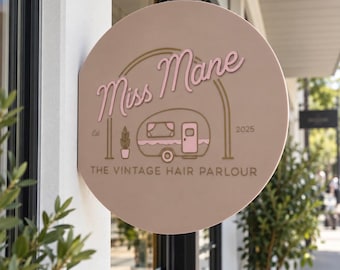 May include: A circular, light tan sign featuring the text Miss Mane in pink script, with a graphic of a small camper, a potted plant, and the text THE VINTAGE HAIR PARLOUR, Est 2025. The sign is mounted on a white wall near a storefront window.