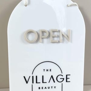 May include: An arched white acrylic sign featuring the word OPEN in raised beige lettering. Below, the text THE VILLAGE BEAUTY is printed in black, framed by a thin black arch line. The sign includes a white rope for hanging at the top.