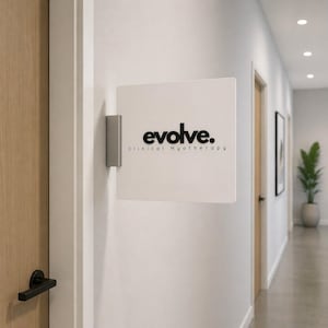Könnte beinhalten: Ein weißes quadratisches Schild mit dem Wort "evolve." in schwarzer Schrift und dem Text "Clinical Myotherapy" darunter. Das Schild ist an einer grauen Metallhalterung an einer weißen Wand befestigt. Das Schild befindet sich in einem Flur mit Holztüren und einer Topfpflanze.