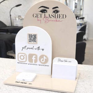 May include: A beige and white acrylic display stand featuring the text GET LASHED by Brooke with an eye graphic. It includes a QR code, social media icons for Instagram, Facebook, and TikTok, a card holder labeled Please take one, and a payment terminal.