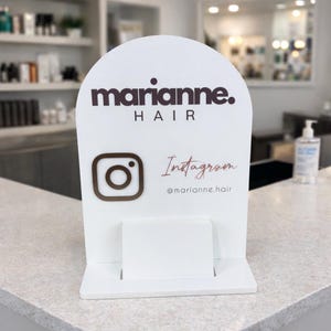 Puede incluir: Un letrero blanco arqueado con el texto "marianne. HAIR" en marrón oscuro. Debajo, un logotipo de Instagram y la palabra "Instagram" en cursiva, con el identificador @marianne.hair. El letrero tiene un pequeño estante en la parte inferior.