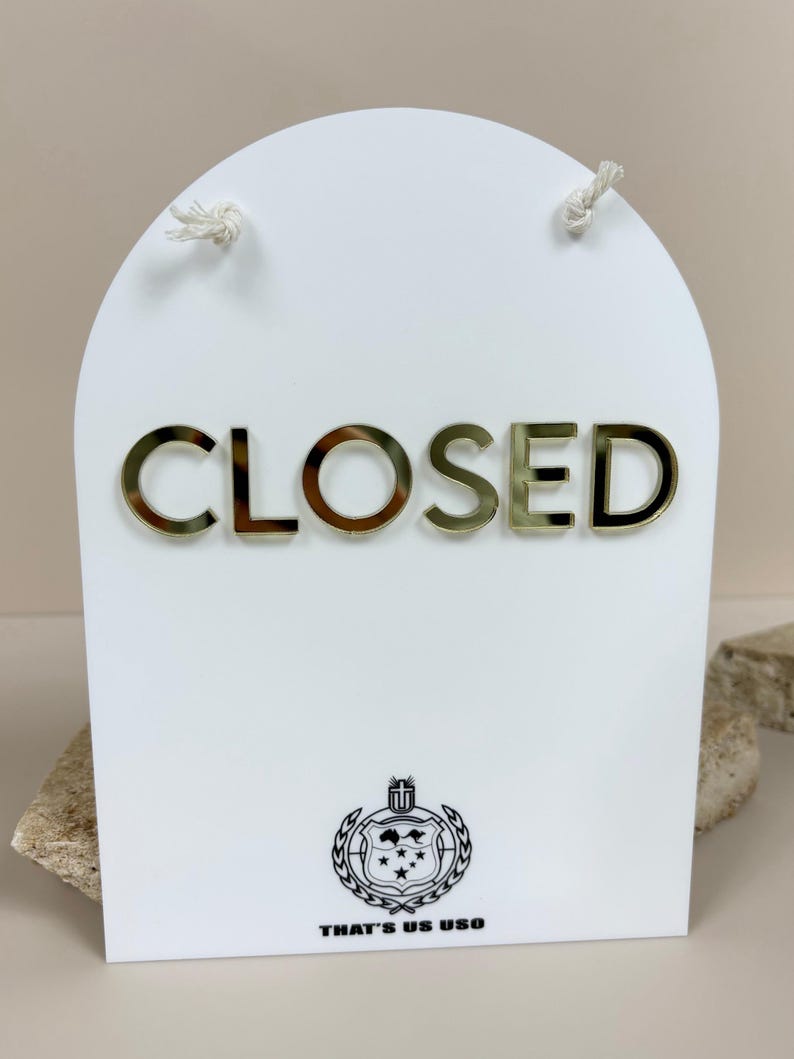 May include: A white arched sign featuring the word CLOSED in gold metallic lettering. At the bottom, a black circular emblem displays a map of Australia with stars and the text THAT'S US USO. Two white strings are attached to the top.