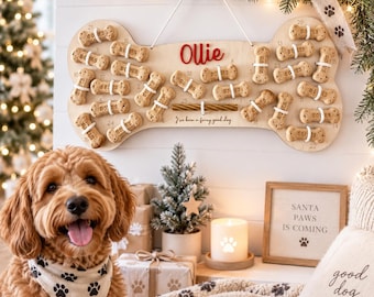 May include: A light-colored wooden dog bone-shaped advent calendar with the name Ollie in red script, featuring twenty-five small bone-shaped treats attached with white elastic loops, and the text I've been a furry good dog printed below.