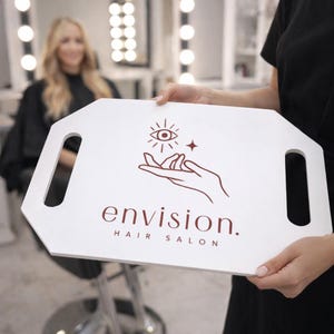 May include: A white rectangular board with two side handles featuring a dark red graphic of a hand holding an eye and a star, with the text "envision. HAIR SALON" printed in dark red below the graphic on the front surface of the board.