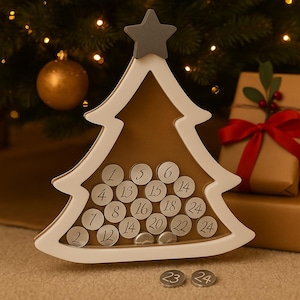 Puede incluir: Un calendario de adviento de madera con forma de árbol de Navidad, con un marco blanco y una estrella gris en la parte superior. El calendario tiene 24 aberturas circulares numeradas. Dos monedas de plata con los números 23 y 24 están en la parte inferior.