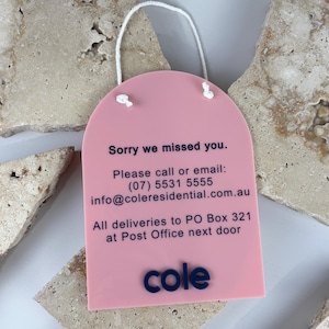 May include: A pink arched acrylic sign with a white hanging cord. The text reads: Sorry we missed you. Please call or email: (07) 5531 5555 info@coleresidential.com.au All deliveries to PO Box 321 at Post Office next door. The word cole is at the bottom.