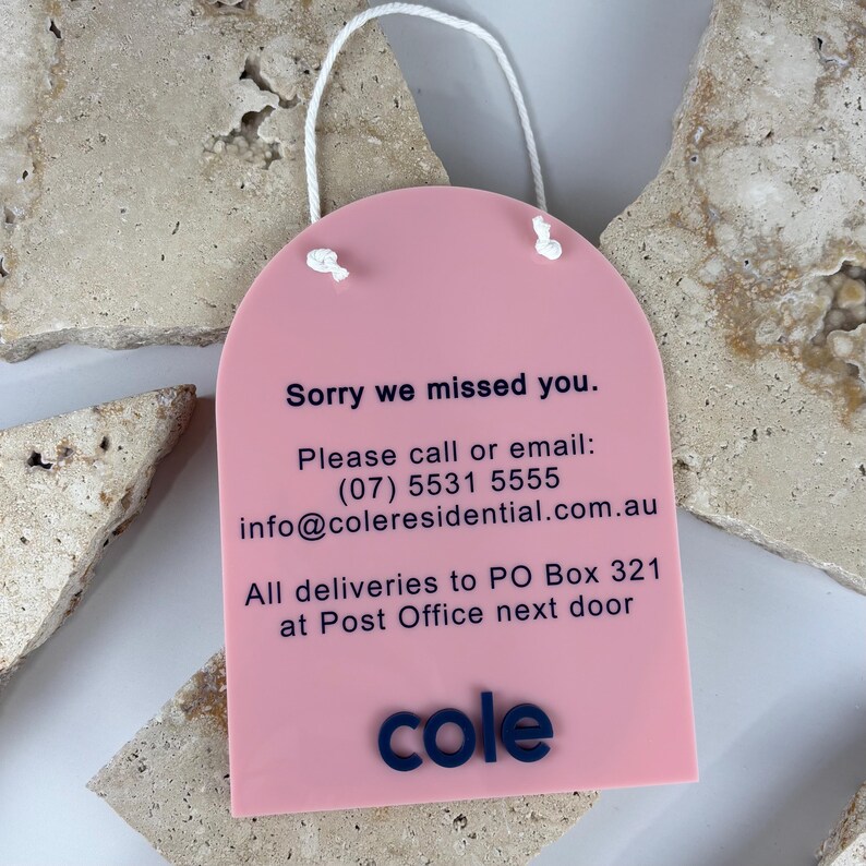 May include: A pink arched acrylic sign with a white hanging cord. The text reads: Sorry we missed you. Please call or email: (07) 5531 5555 info@coleresidential.com.au All deliveries to PO Box 321 at Post Office next door. The word cole is at the bottom.