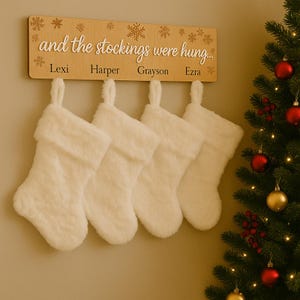 May include: A wooden sign saying "and the stockings were hung..." with names Lexi, Harper, Grayson, and Ezra. Four white, fluffy Christmas stockings hang beneath the sign. A decorated Christmas tree is on the right side of the image.