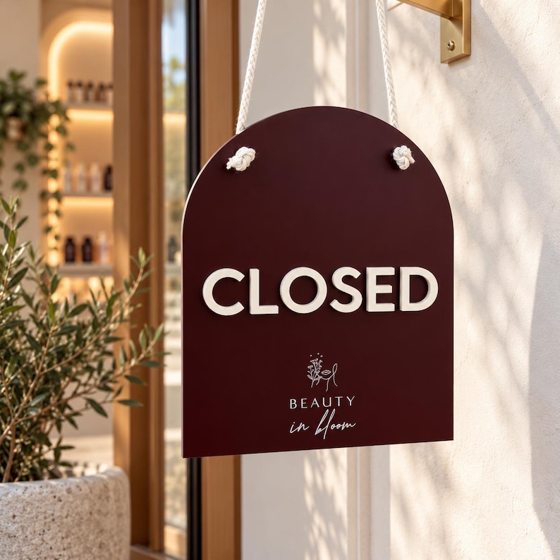May include: A dark maroon arched sign hanging by white ropes. The sign features the word CLOSED in large white capital letters, with the text Beauty in bloom and a line-art floral face illustration printed in white below the main text.