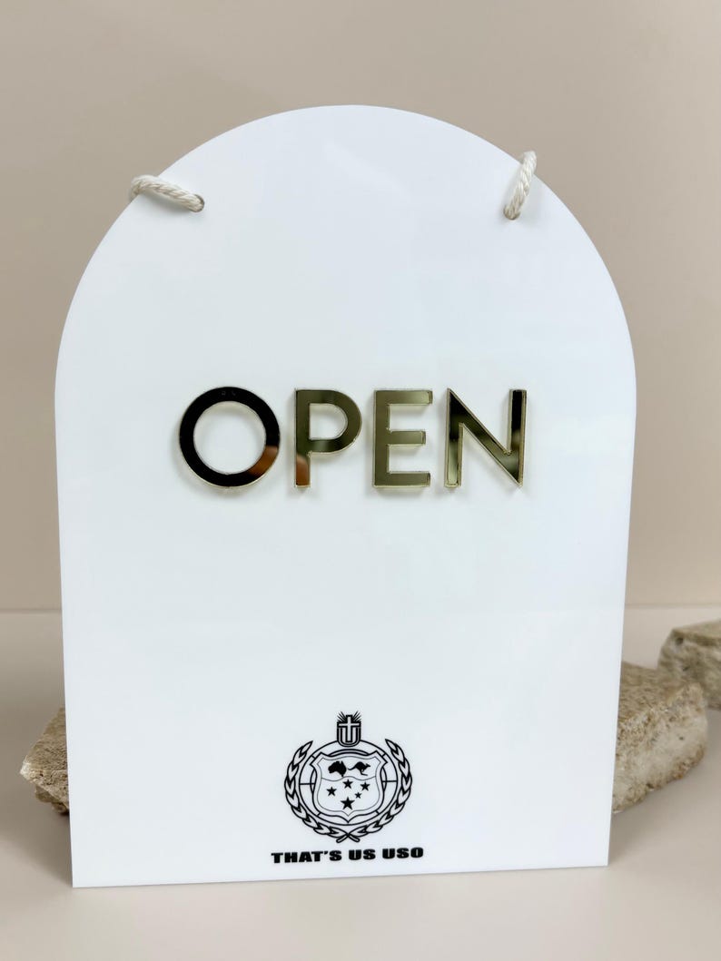 May include: An arched white acrylic sign featuring the word OPEN in gold reflective lettering. Below the text is a black circular emblem with a shield design and the text THAT'S US USO. The sign includes a white rope for hanging.
