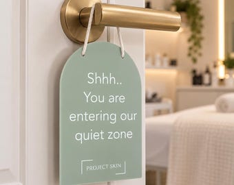 May include: A sage green arched door sign hanging from a brushed gold door handle by a white cord. The sign features white text that reads: Shhh.. You are entering our quiet zone, with the text PROJECT SKIN enclosed in a white bracket design.